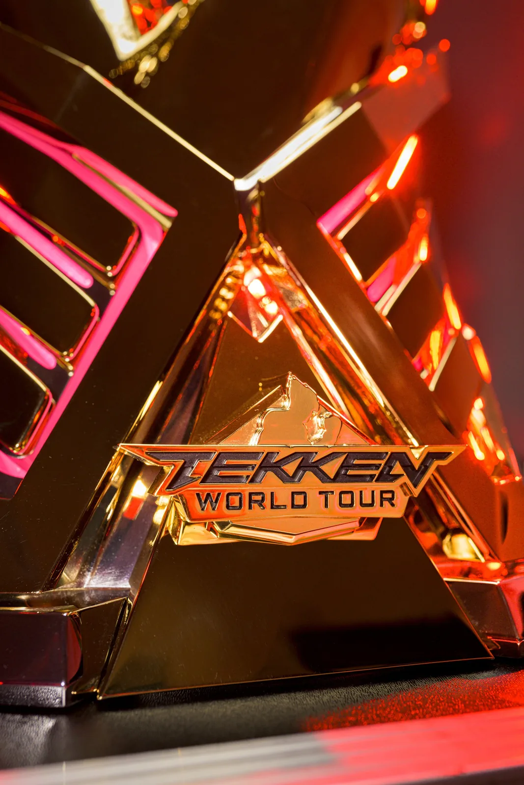 Close view of the finished TEKKEN World Tour 2025 trophy, pink and orange light glowing through the chevrons around the logo plate