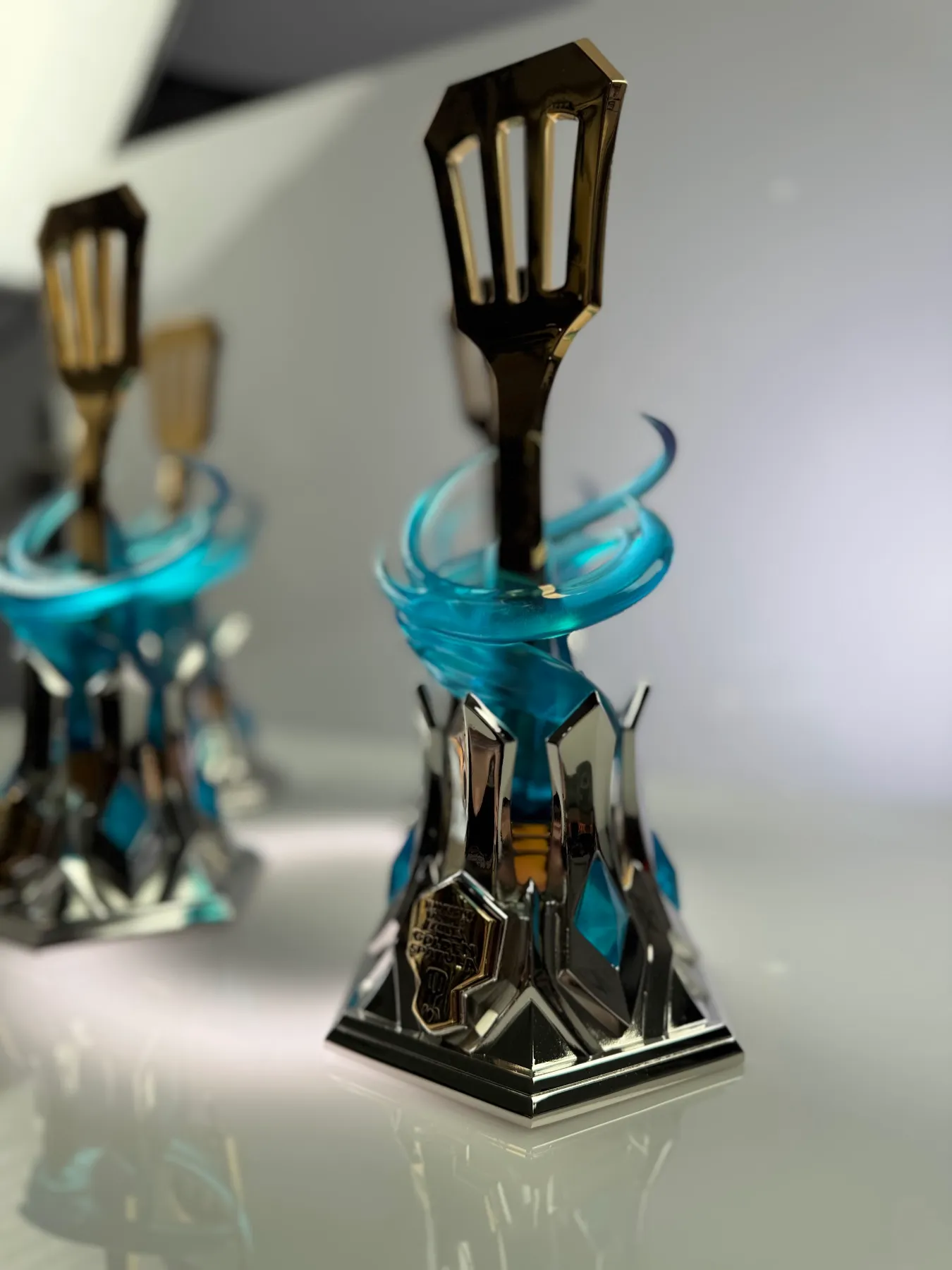 A Teamfight Tactics Golden Spatula trophy built by Hoku Props, a mirror-gold spatula rising through a translucent blue vortex on a chromed hextech base, lit on a white sweep
