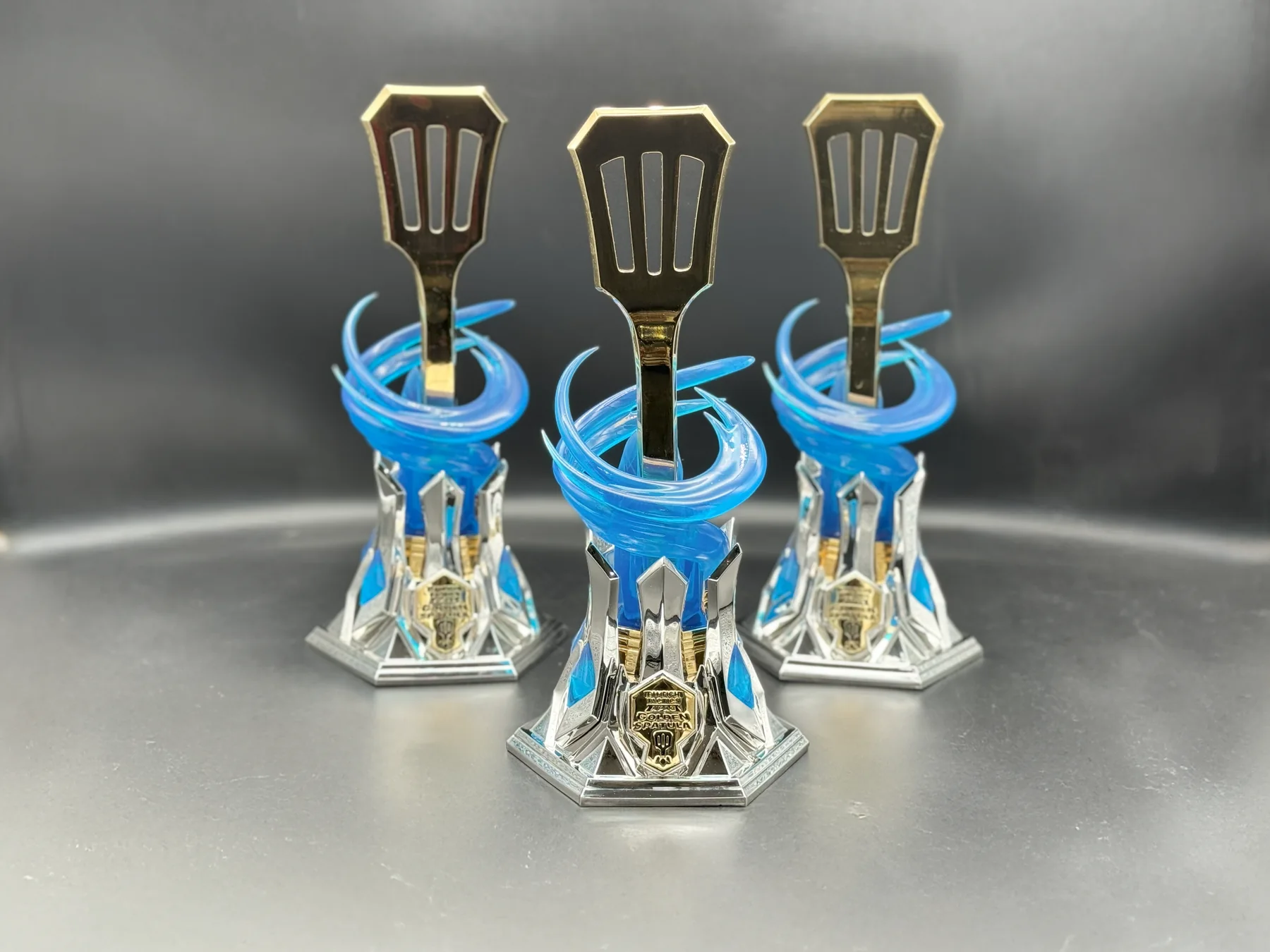 A Teamfight Tactics Golden Spatula trophy built by Hoku Props, a mirror-gold spatula rising through a translucent blue vortex on a chromed hextech base, lit on a white sweep
