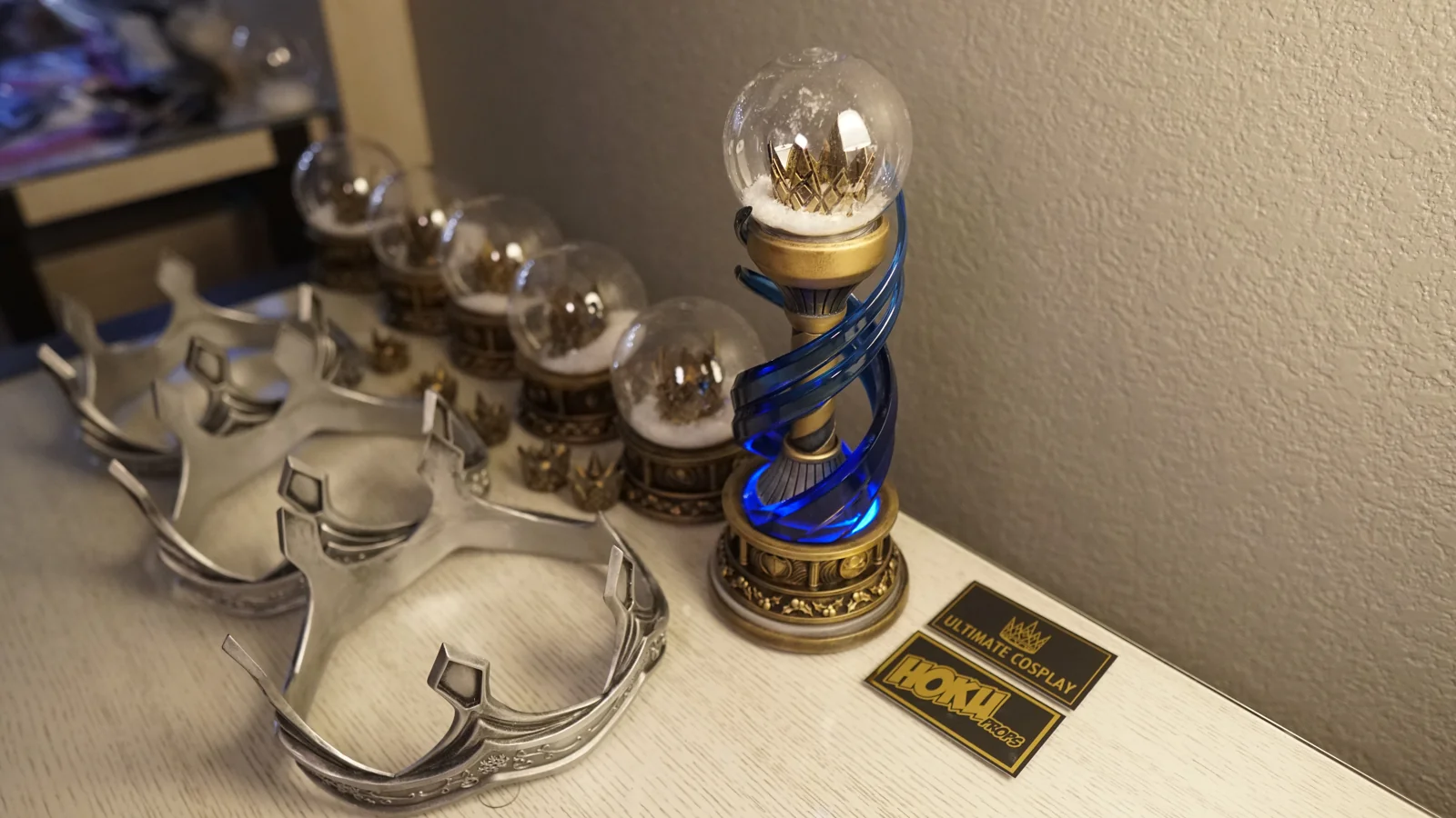 The full Ultimate Cosplay x Holiday Matsuri 2021 award set by Hoku Props: silver crowns, a row of crown snow globes and the lit winner trophy